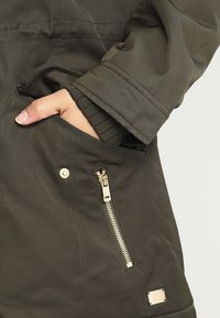 Tmavě zelená látková bunda se zipovou kapsou, s zlatými kovovými detaily a strukturovaným manžetem. Hladký povrch s mírně polstrovaným designem.