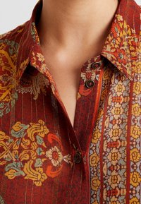Gros plan d'une personne portant une chemise rouge à motifs floraux jaunes et verts avec des boutons foncés près du col.