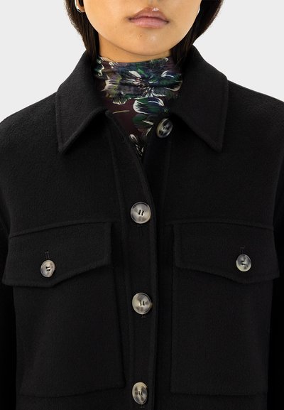 Schwarze Wolle-Mischung Jacke mit einem Kragen-Halsauschnitt, zwei Brusttaschen und sechs Knöpfen. Der Stoff hat eine glatte Textur. Subtiler Glanz auf den Knöpfen.