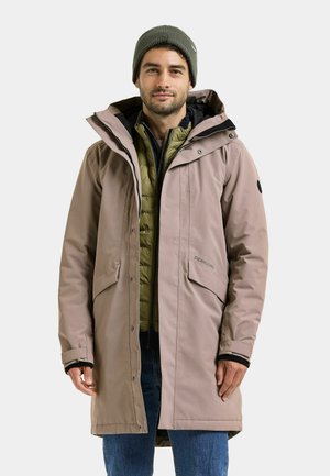 Mann trägt einen beigen langen Wintermantel über einer olivgrünen gesteppte Jacke, blaue Jeans und eine olivgrüne Mütze, schaut nach vorne.