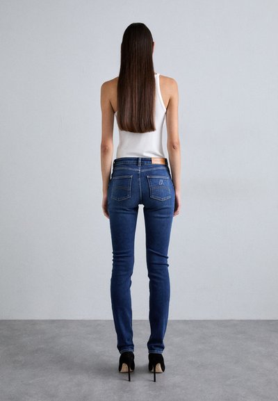 Slim-fit mörkblå jeans med bakfickor och en tan läderlapp. Bärs med en vit linne och svarta högklackade skor. Grå flat bakgrund.