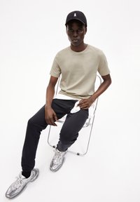 Beige t-shirt, donkere trainingsbroek en grijze sneakers. Model zit op een doorzichtige stoel. Zwarte pet met een wit logo. Eenvoudig, modern ontwerp.