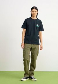 Camiseta de algodón negra con gráfico verde en el pecho, combinada con pantalones cargo holgados en color caqui con múltiples bolsillos y zapatillas negras.