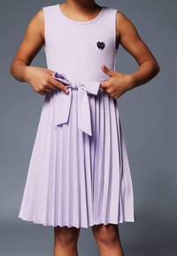 Dziecko w bezrękawowej lawendowej sukience plisowanej, zawiązujące materiałową kokardę w pasie, z małą łatką w kształcie serca na klatce piersiowej.