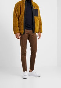 Faux fur gul jacka med en svart dragkedjeficka, tillsammans med bruna byxor och vita sneakers. Jackan har en hög krage och en avslappnad passform.