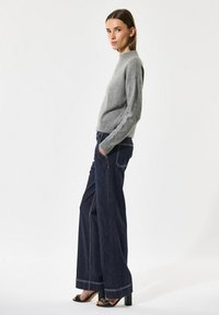 Sieviete pelēkā garā piedurkņu džemperī un tumšās platas biksēs stāv sāniski, rokas ieliktas kabatās, pret baltu fonu.