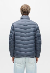 Sötétkék párnázott kabát, chevron steppelésű dizájnnal, magas gallérral és sima felülettel. Világos színű farmer felett viselve.