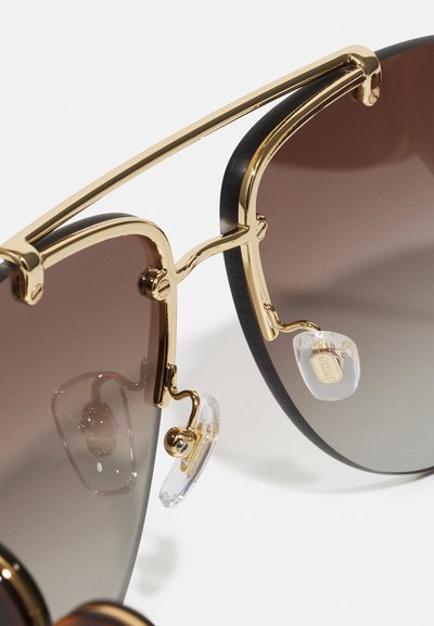 Oversized sunglasses with gradient brown lenses. Gold metal accents and clear nose pads. Black frame with a smooth finish.