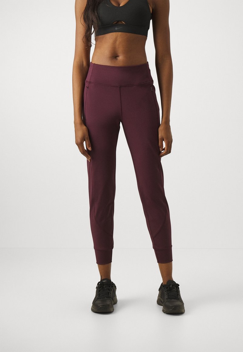 Femme portant un soutien-gorge de sport noir, un pantalon de jogging taille haute bordeaux avec poches zippées, et des baskets noires, se tenant devant un fond uni.