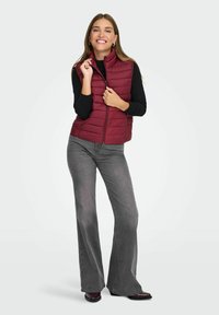 Burgundy puffer väst med uppstående krage och dragkedja framtill, bärs över en svart långärmad tröja. Grå utsvängda jeans och svarta skor fulländar looken.