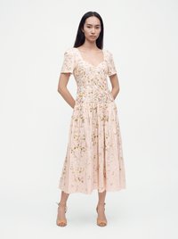 Kvinde iført en lyserød, midilængde kjole med blomsterbroderi, korte pufærmer og beige højhælede sandaler, stående med hænderne bag ryggen.