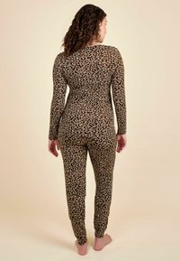Žena s kučeravými vlasmi, oblečená v priliehavom dlhom rukáve s leopardím vzorom a zodpovedajúcich nohaviciach, stojí naboso s chrbtom otočeným k bezvýraznému pozadiu.