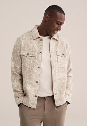 Glimlachende man die een gestructureerd beige jasje draagt over een licht hemd en een bruine broek, handen in de zakken, kijkt naar beneden naar rechts.