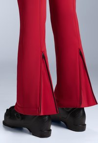 Piros elasztikus anyagból készült sínadrág, oldalán cipzárokkal a boka résznél. Fekete síbakancsokkal viselve, elegáns kialakítást mutatva.