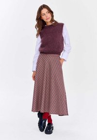 Burgundská chlupatá bezrukávová svetr přes bílou košili, sladěná s červenou a černou kostkovanou sukní. Modelka má na sobě černé platformové boty a červené ponožky.