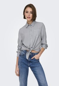 Camicia a righe blu e bianca, annodata in vita. Collo classico, maniche lunghe, taschino sul petto, abbinata a jeans blu.