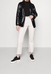 Schwarze Kunstleder-Blazer, geripptes schwarzes Oberteil, weiße gerade Jeans und klobige schwarze Sneakers mit weißen Sohlen. Einfaches und modernes Ensemble.