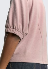 Pull rose court avec des manches bouffantes, des poignets froncés et une texture douce. Détail de couture visible sur le côté. Porté par-dessus un denim foncé.