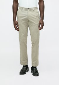 Beige Chinos mit geradem Schnitt und Knopfverschluss. Glatter Baumwollstoff, mit Seitentaschen und umgeschlagenem Saum. Kombiniert mit schwarzen Loafers.