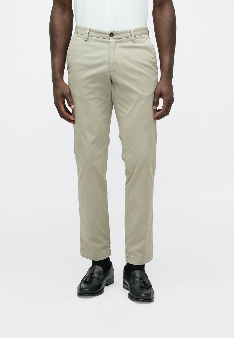 Beige Chinos mit geradem Schnitt und Knopfverschluss. Glatter Baumwollstoff, mit Seitentaschen und umgeschlagenem Saum. Kombiniert mit schwarzen Loafers.