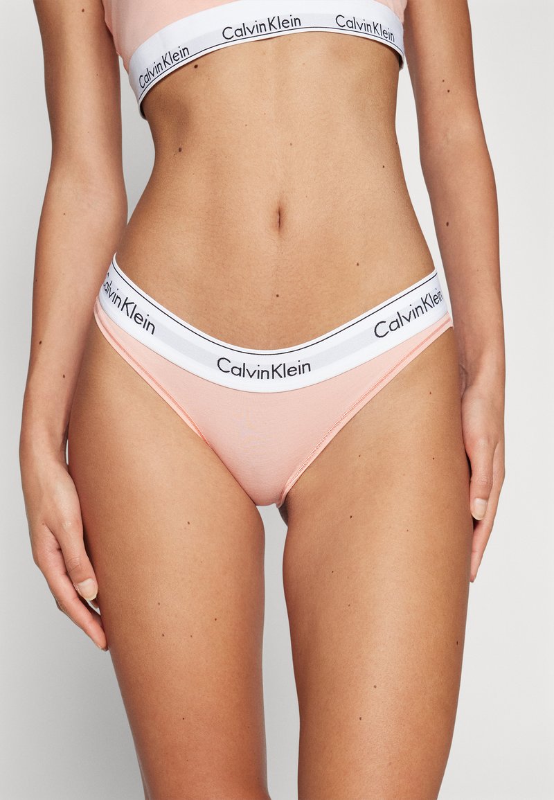 Zestaw bielizny damskiej Calvin Klein wykonany z miękkiego brzoskwiniowego materiału, z białym elastycznym paskiem w talii z czarnym logo, zaprojektowany z myślą o komforcie i dopasowaniu.