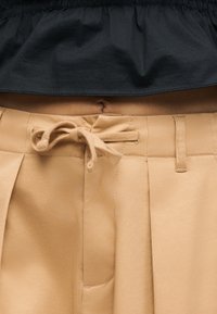 Κοντινό πλάνο στις μπεζ παντελόνες με δεμένη υφασμάτινη ζώνη, θηλιές για ζώνη και μαύρη ελαστική μέση πάνω από τον ομφαλό.