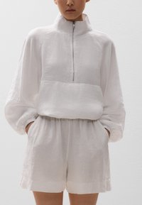 Weißes Leinenoberteil mit hohem Kragen und halbem Reißverschluss, kombiniert mit passenden Shorts. Hat eine lockere Passform und elastische Bündchen für den Komfort.