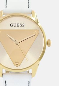 Gouden driehoekige wijzerplaat met minimalistische wijzers, voorzien van een witte leren band met zwarte stiksels en het "GUESS" logo.