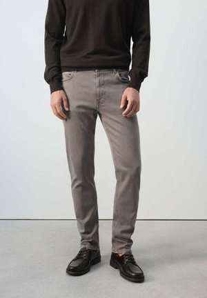 Mann trägt graue Slim-Fit-Jeans, ein schwarzes Langarmshirt und schwarze Lederschuhe und steht vor einem schlichten Hintergrund.