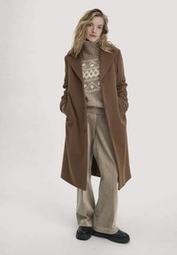 Brauner Wollmantel mit weitem Revers, kombiniert mit einem hellen, gemusterten Pullover und beigen Cordhosen. Schwarze Plateauschuhe vervollständigen das Outfit.