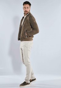 Braune Lederbomberjacke mit gerippten Bündchen, Reißverschluss vorne und Seitentaschen; kombiniert mit hellbeigen Hosen und dunkelbraunen Sneakers.