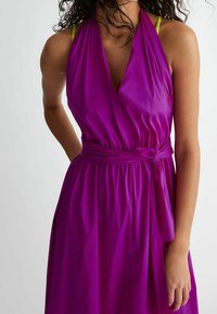 Fioletowa sukienka na ramiączkach z lekkiego materiału, z głębokim dekoltem w kształcie litery V, detalem w postaci wiązania w talii oraz swobodnym fasonem. Widoczne kontrastowe limonkowe ramiączka.