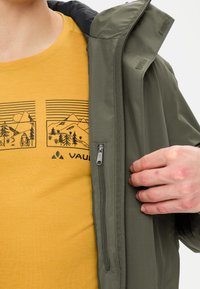 Giacca verde oliva con tasca con zip, indossata sopra una maglietta gialla con grafica che mostra illustrazioni di montagne e dettagli neri.