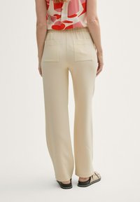 Pantalone krem boje s teksturiranom površinom, elastičnim strukom i dva strašnja džepa. Model nosi crne cipele bez pete.