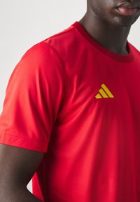 Röd idrottströja med korta ärmar, utrustad med en gul tre-stripes-logotyp på vänster bröstområde. Slät, lätt tyg.