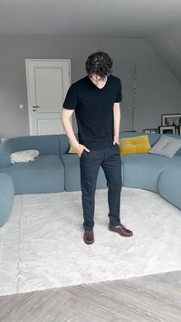 Černá košile s krátkým rukávem, námořnické kalhoty a hnědé boty. Stojí na světlém koberci v moderní místnosti s modrou pohovkou a dekoracemi.