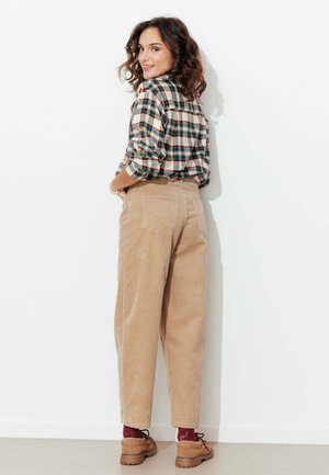 Femme aux cheveux bouclés, portant un pantalon en velours côtelé beige, une chemise à carreaux et des chaussures marron, se tenant de dos, regardant par-dessus son épaule et souriant devant un mur blanc.