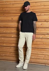 Černé bavlněné tričko, béžové bavlněné kalhoty, bílé tenisky. Osoba má tetování a nosí černou kšiltovku. Pozadí ze dřeva.