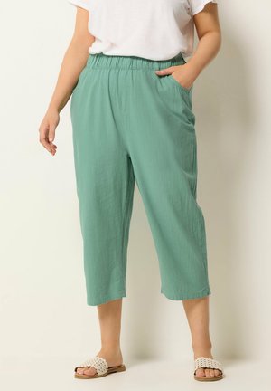 Osoba ubrana w zielone, krótkie spodnie z elastycznym pasem i bocznymi kieszeniami, w połączeniu z białymi, szydełkowanymi klapkami i białą górą.