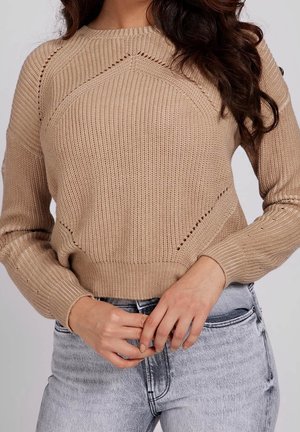 Femme portant un pull beige en maille avec des motifs géométriques et un jean gris clair délavé, mains jointes à la taille, sur fond uni.