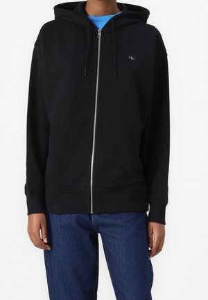 Persona que lleva una sudadera negra con cremallera y un pequeño logo en el pecho y vaqueros azules, de pie frente a un fondo blanco liso.