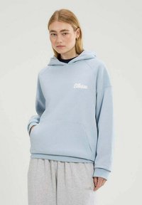 Sudadera azul claro con un bolsillo frontal y logotipo blanco, con mangas raglán y puños de canalé, combinada con pantalones de chándal grises claros.