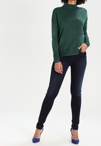 Pull en tricot vert à manches longues et col montant, assorti à un jean slim foncé et des chaussures à talons bleues.