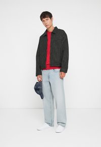 Chaqueta a rayas blanco y negro, camiseta roja, vaqueros de color azul claro, zapatillas blancas y una gorra azul en la mano, de pie contra un fondo blanco.