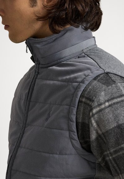 Grå polstret vest med høj krave, der har vandrette syninger og et logo. Kombineret med en grå ternet skjorte med lange ærmer under.