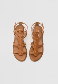 Sandales gladiateur en cuir marron avec plusieurs brides, semelle plate et fermeture à boucle. Texture lisse et design minimaliste.