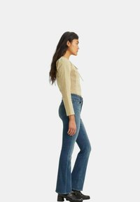 Jeans de mezclilla acampanados en azul oscuro combinados con una blusa ajustada y texturizada en beige claro que presenta un diseño recogido y un detalle de lazo en el escote.