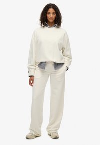 Sudadera de crema con hombros caídos superpuesta sobre una camisa de cuadros azul con cuello, combinada con pantalones de tiro ancho en crema a juego y zapatillas beige.