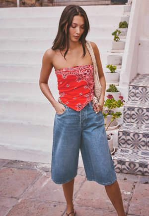 Woman wearing red patterned tube top, long denim shorts, and carrying a woven shoulder bag, standing near white stairs and tiled steps with plants.