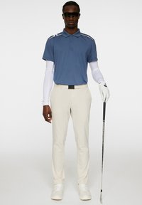 Vaaleansininen poolopaita, valkoiset pitkät hihat alla, vaaleat housut ja valkoiset lenkkarit. Pitää golfmailaa mustalla kädensijalla. Mustat aurinkolasit.